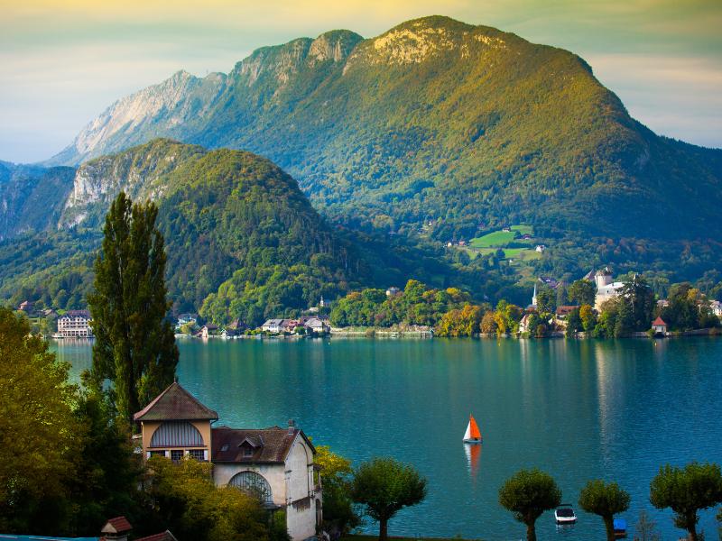 Consultant SEO Annecy - Vue du lac d'Annecy entouré de montagnes avec une petite voile rouge sur l'eau.