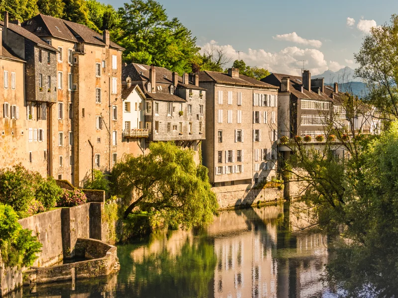 consultant seo oloron - Vue pittoresque des bâtiments historiques d'Oloron-Sainte-Marie, bordant une rivière entourée de verdure sous une lumière dorée.